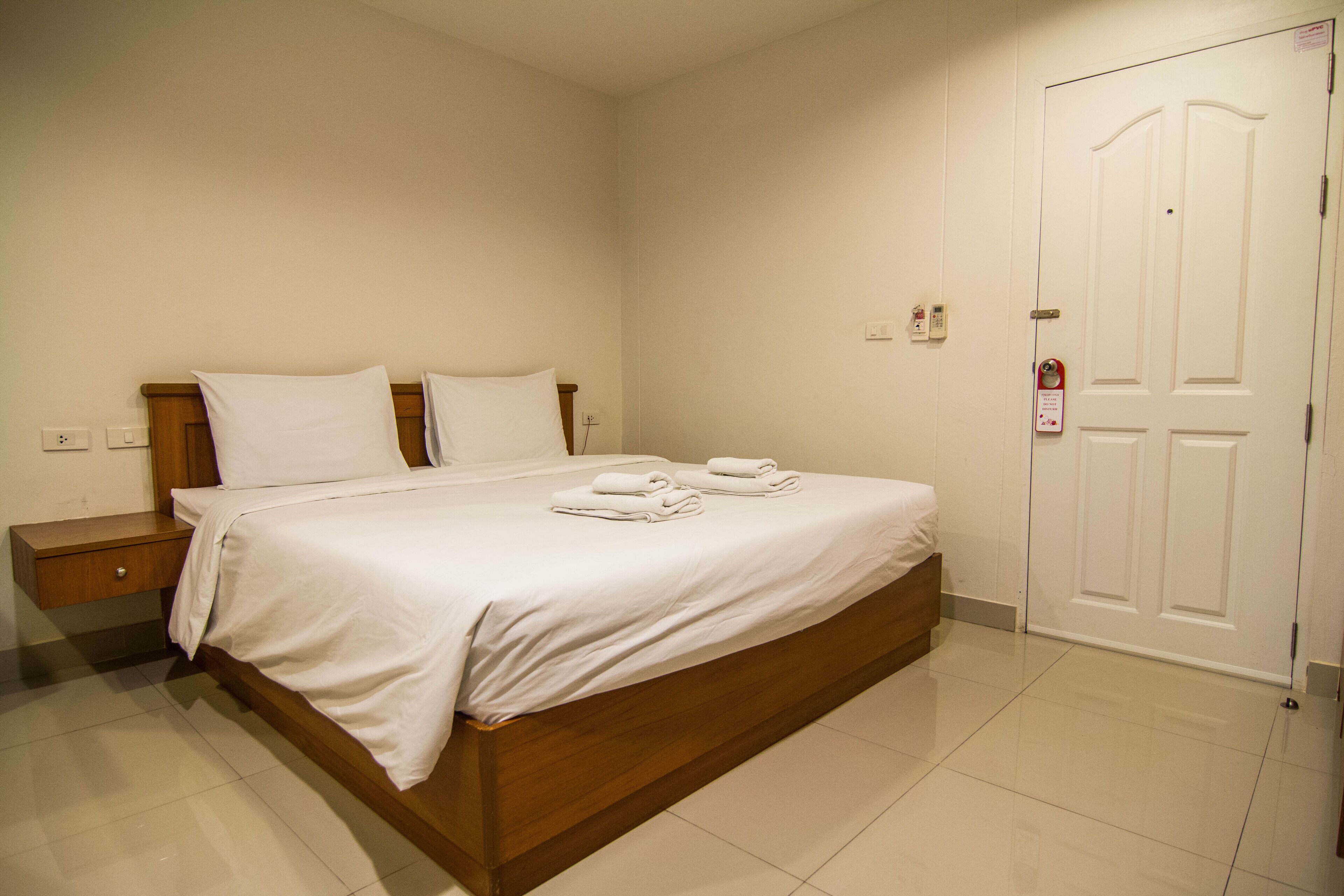 Una cama bien hecha con sábanas y toallas blancas, cabecera de madera, mesita de noche y puerta blanca con cerradura.