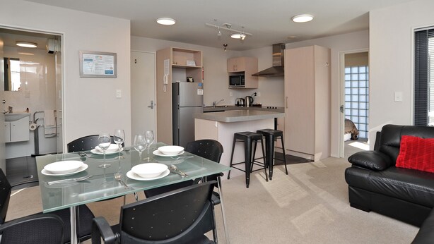 A modern kitchen with a microwave, refrigerator, and gas stove. There is a dining area with a glass table and black chairs. A black leather sofa is visible in the living room area. - Carlton Villa 3 (Christchurch)