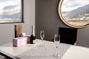 Ein Tisch mit einer Champagnerflasche, zwei Gläsern und einer pinken Schachtel, vor einer Berg- und Stadtlandschaft.