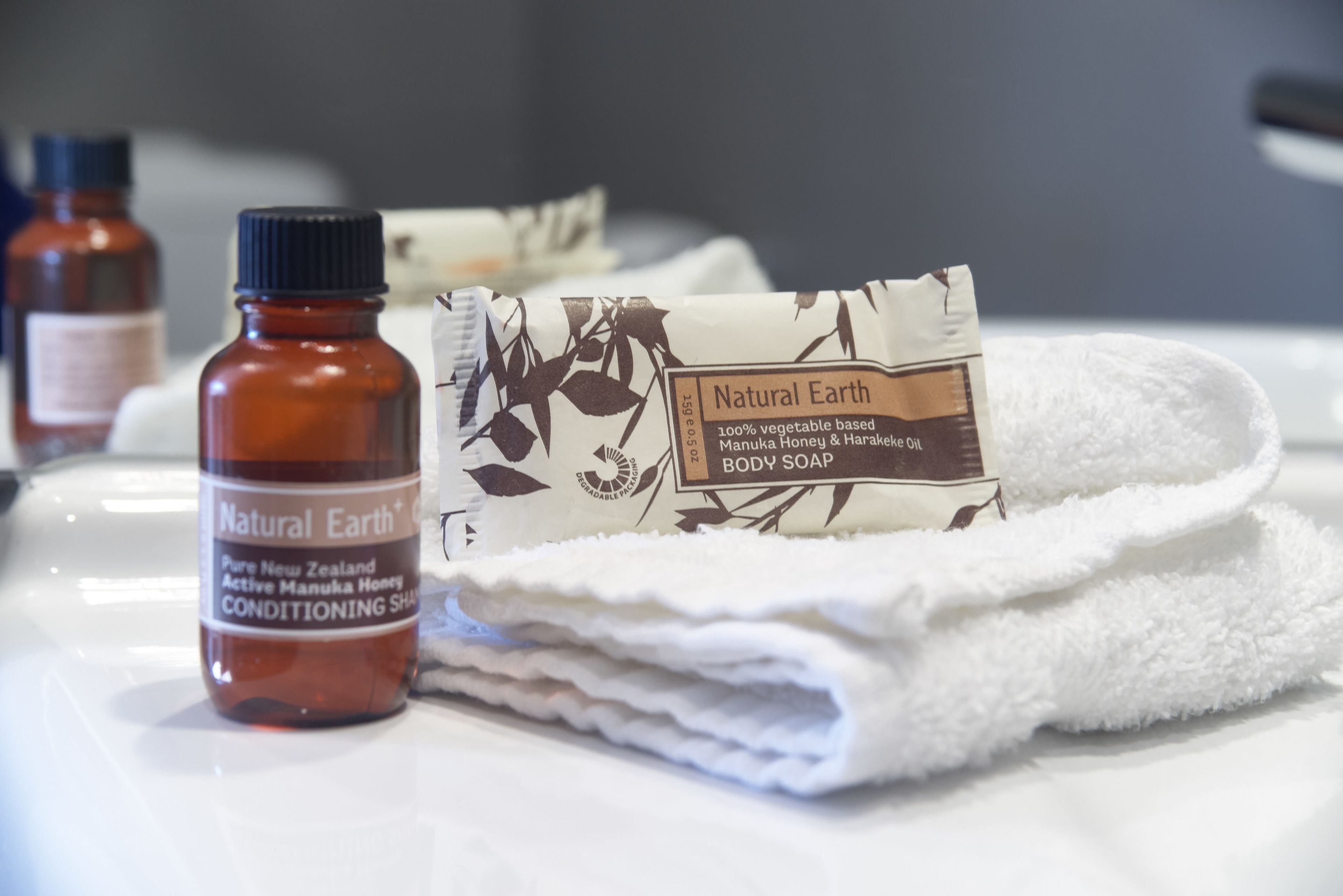 A bathroom with a bottle of Natural Earth conditioning shampoo, a bar of Manuka Honey & Harakeke body soap, and a folded white towel.