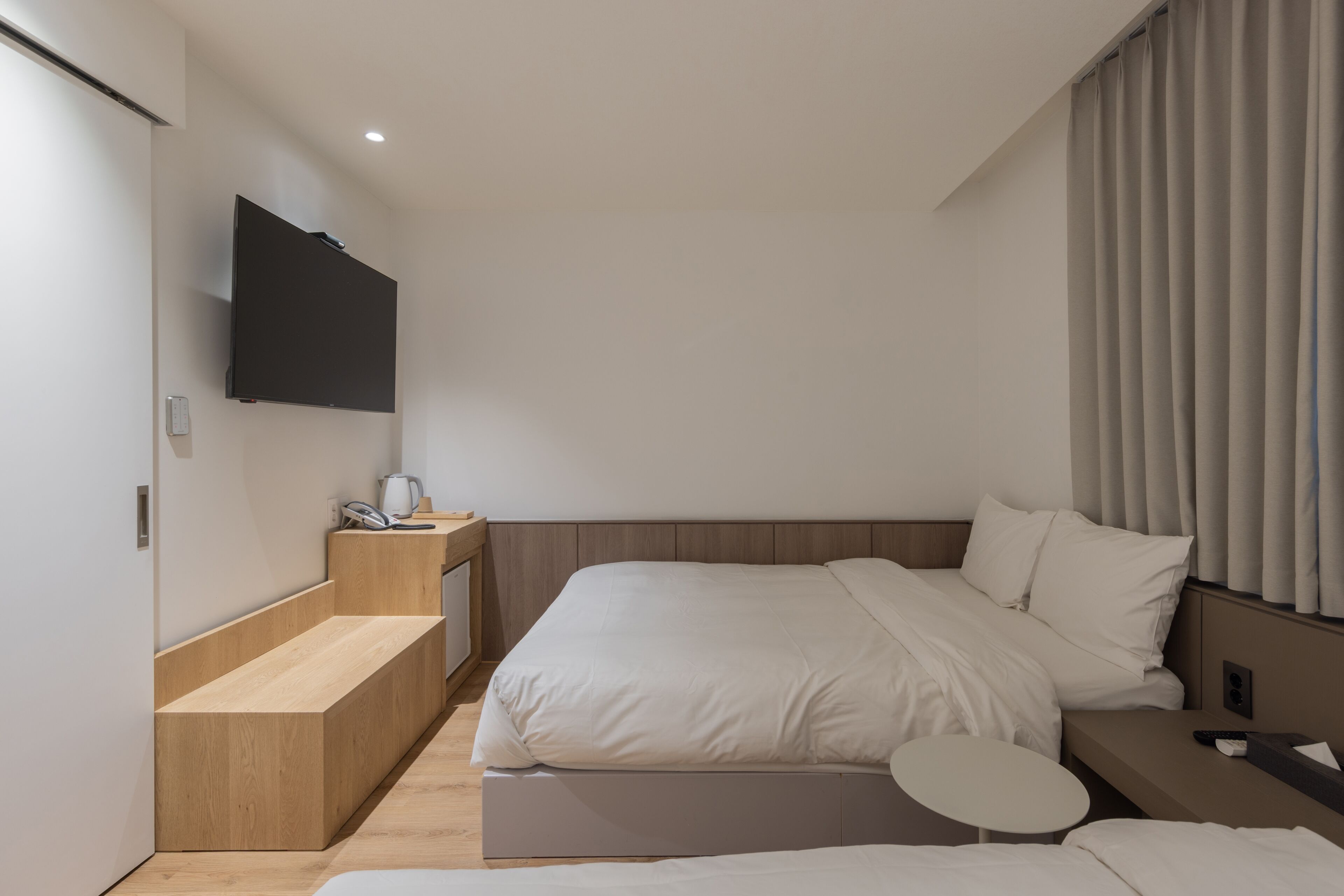 A modern hotel room with a large bed, a bedside table, a small round table, and a mounted TV.