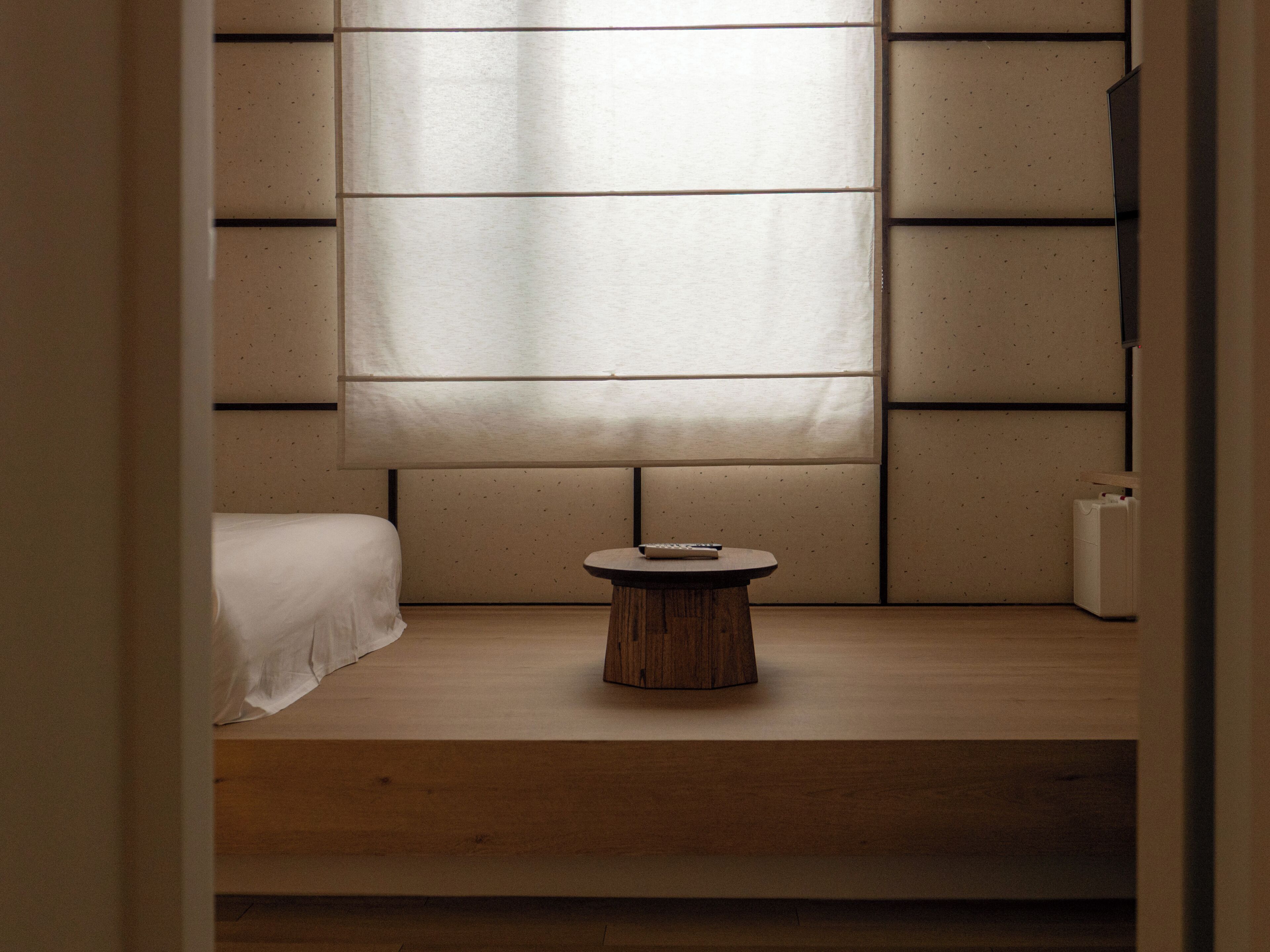 Una habitación minimalista con una cama, una pequeña mesa de madera y un ventanal grande.