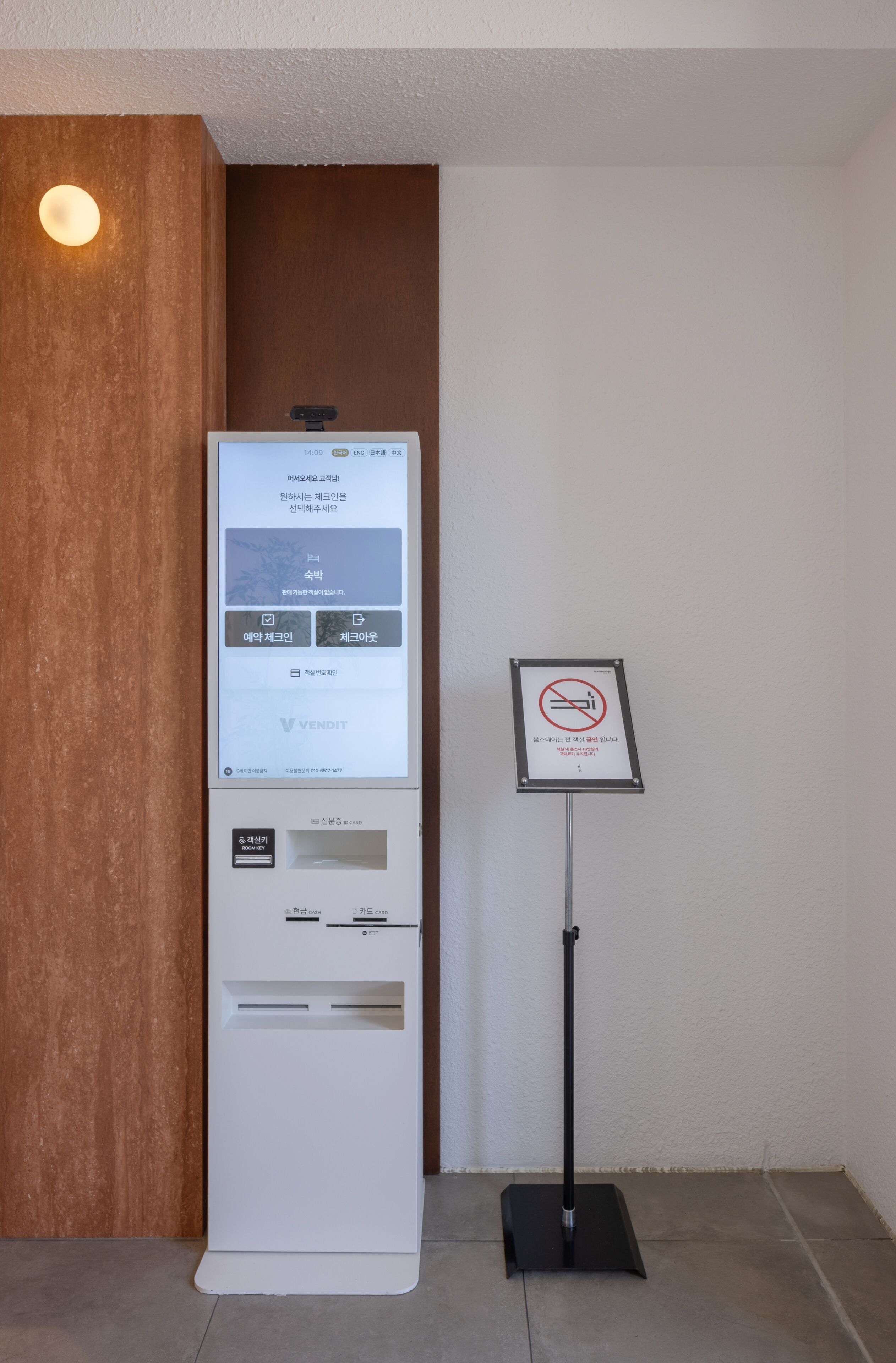 Un quiosco de autoservicio con pantalla digital, un letrero de prohibición de fumar y un soporte metálico.