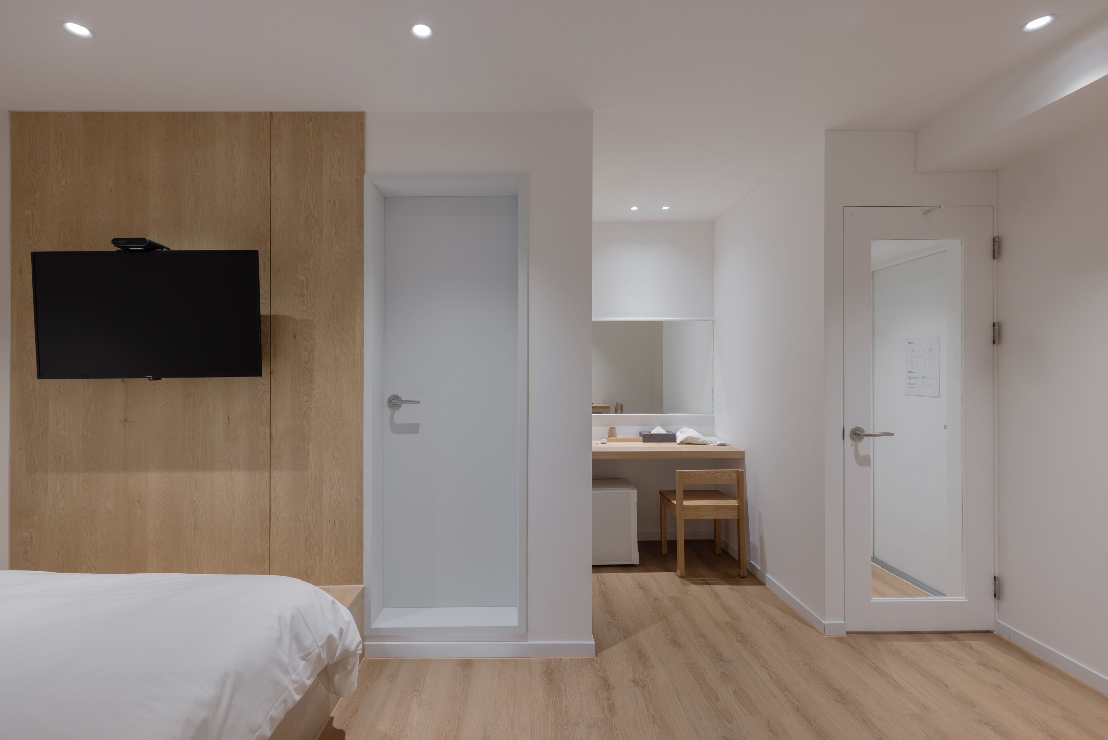 A bedroom with a bed, a TV mounted on a wooden panel, a desk with a chair, and a bathroom with a mirror and door.