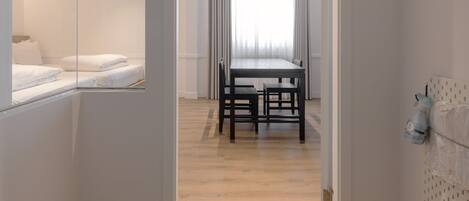 A hotel room with a wooden floor, a bed with white bedding, a dining area with a table and chairs, and a mirror on the wall.