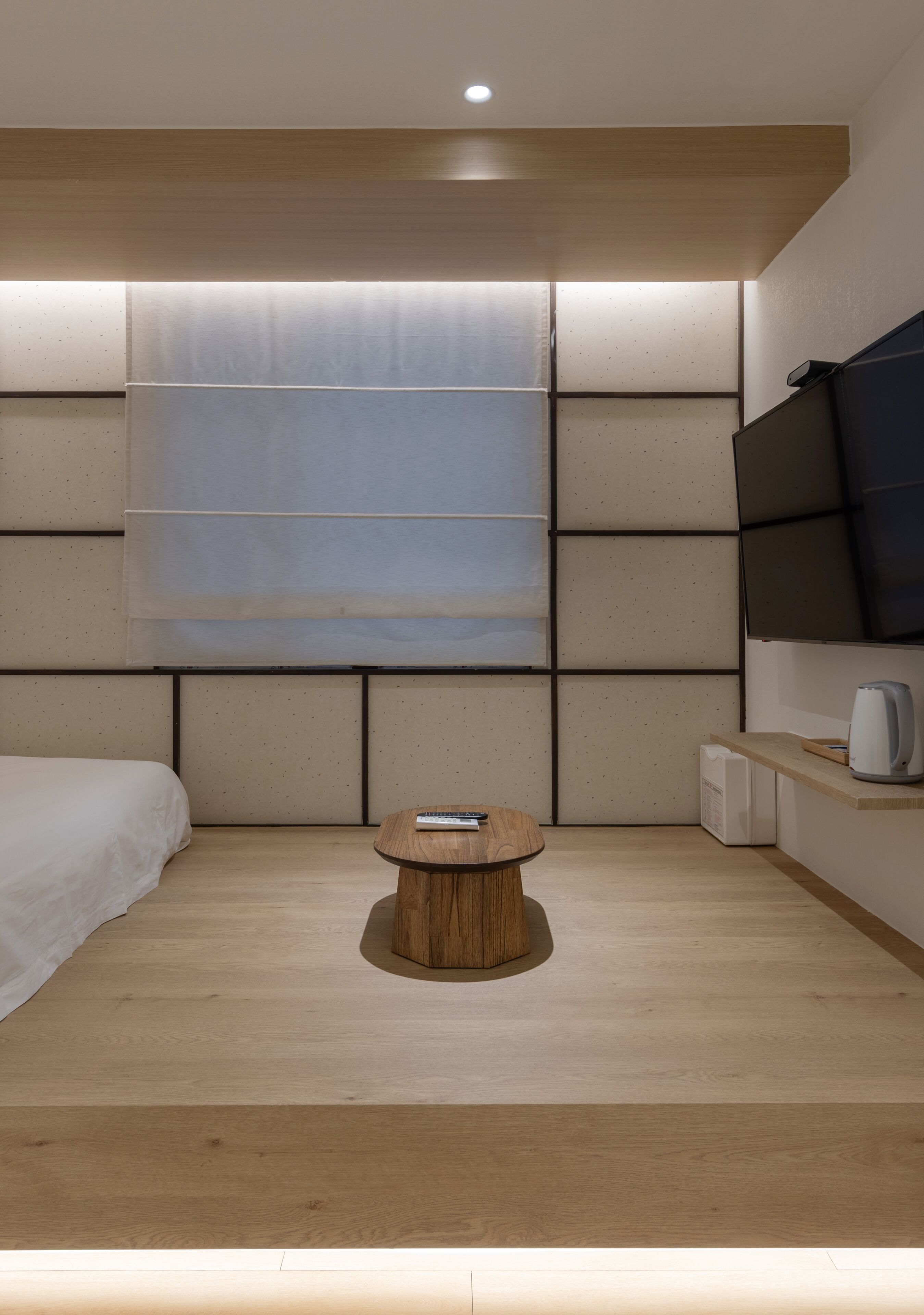 Un dormitorio moderno con una cama, una mesa redonda de madera, un televisor en la pared y una ventana con persianas.