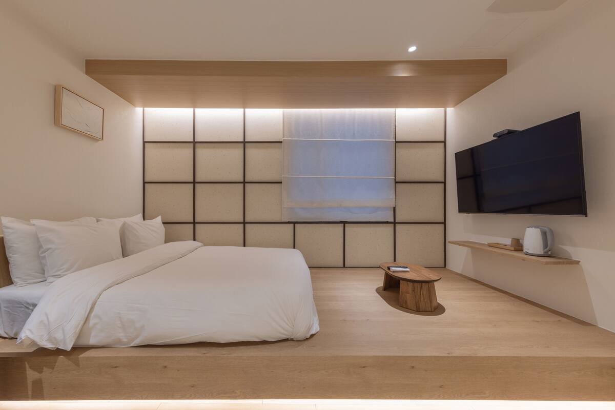 A modern bedroom with a bed, a wooden coffee table, a TV mounted on the wall, and a window with blinds.