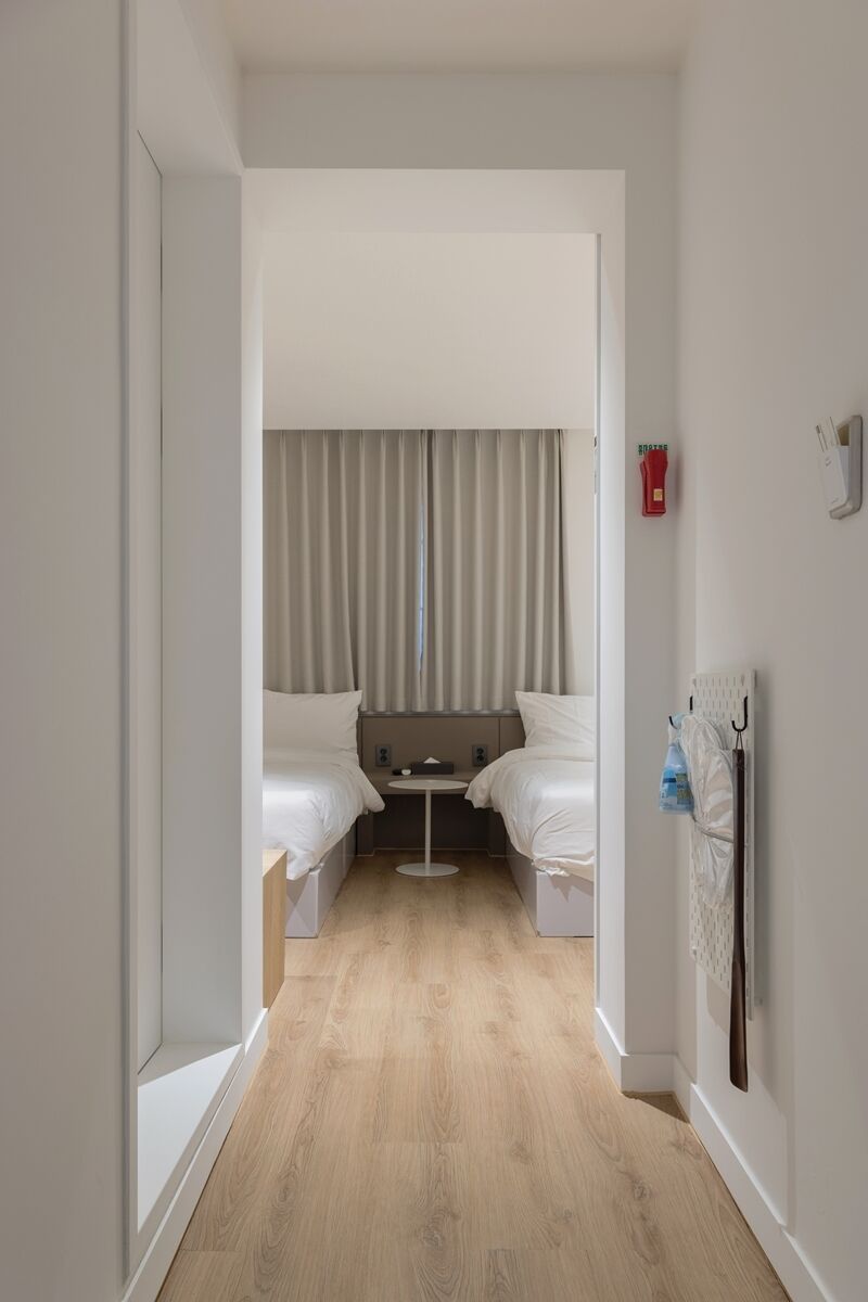 A narrow hallway leading to a room with two beds, a small table, and a window with curtains.