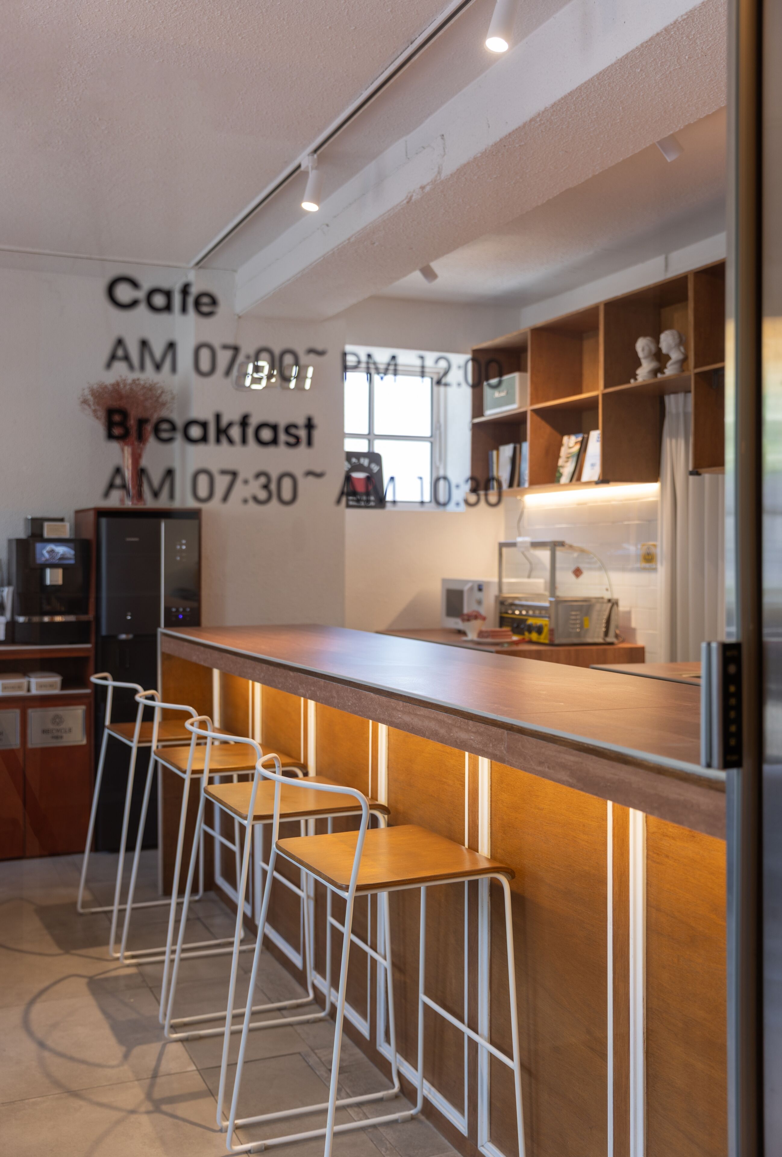 A cafe with a wooden counter, bar stools, a refrigerator, and a wall clock displaying cafe hours.