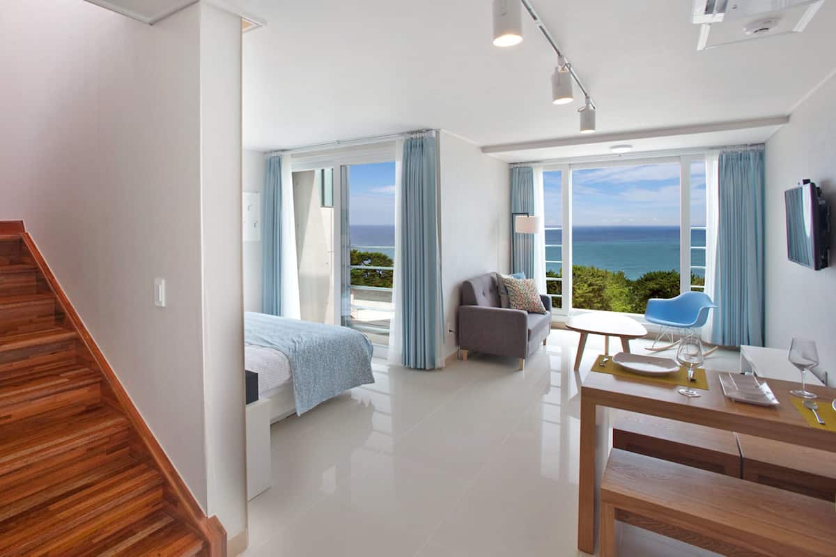 A modern hotel room with a wooden staircase, a bed, a balcony with a view of the sea, a dining area with a table and chairs, and a flat-screen TV mounted on the wall.