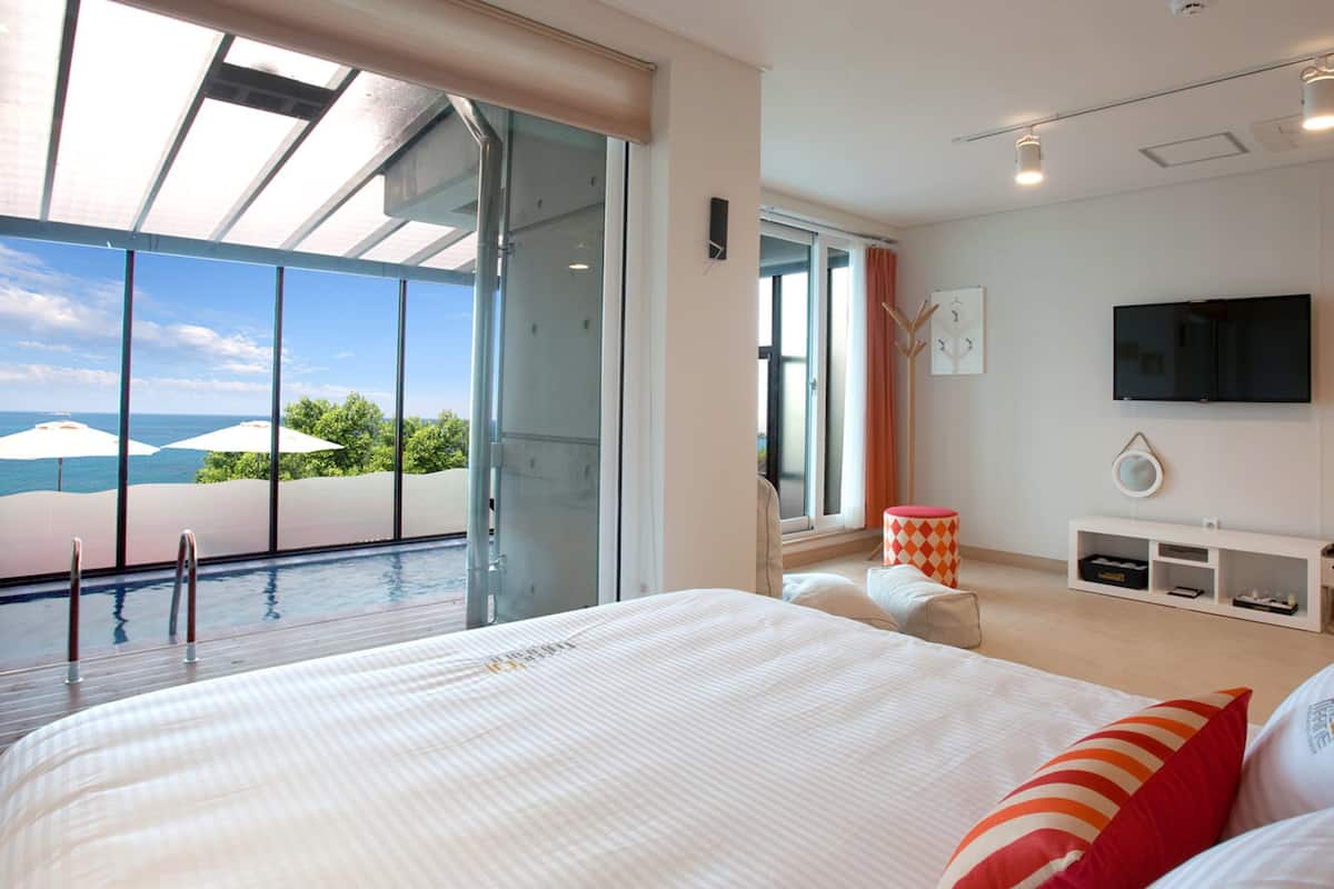 A modern hotel room with a large bed, a TV, and a view of the ocean through large windows.