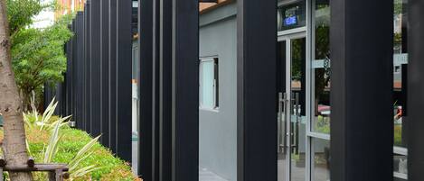 Un bâtiment moderne doté d’une entrée en verre, de colonnes noires verticales et d’un jardin bien entretenu avec un banc en bois.