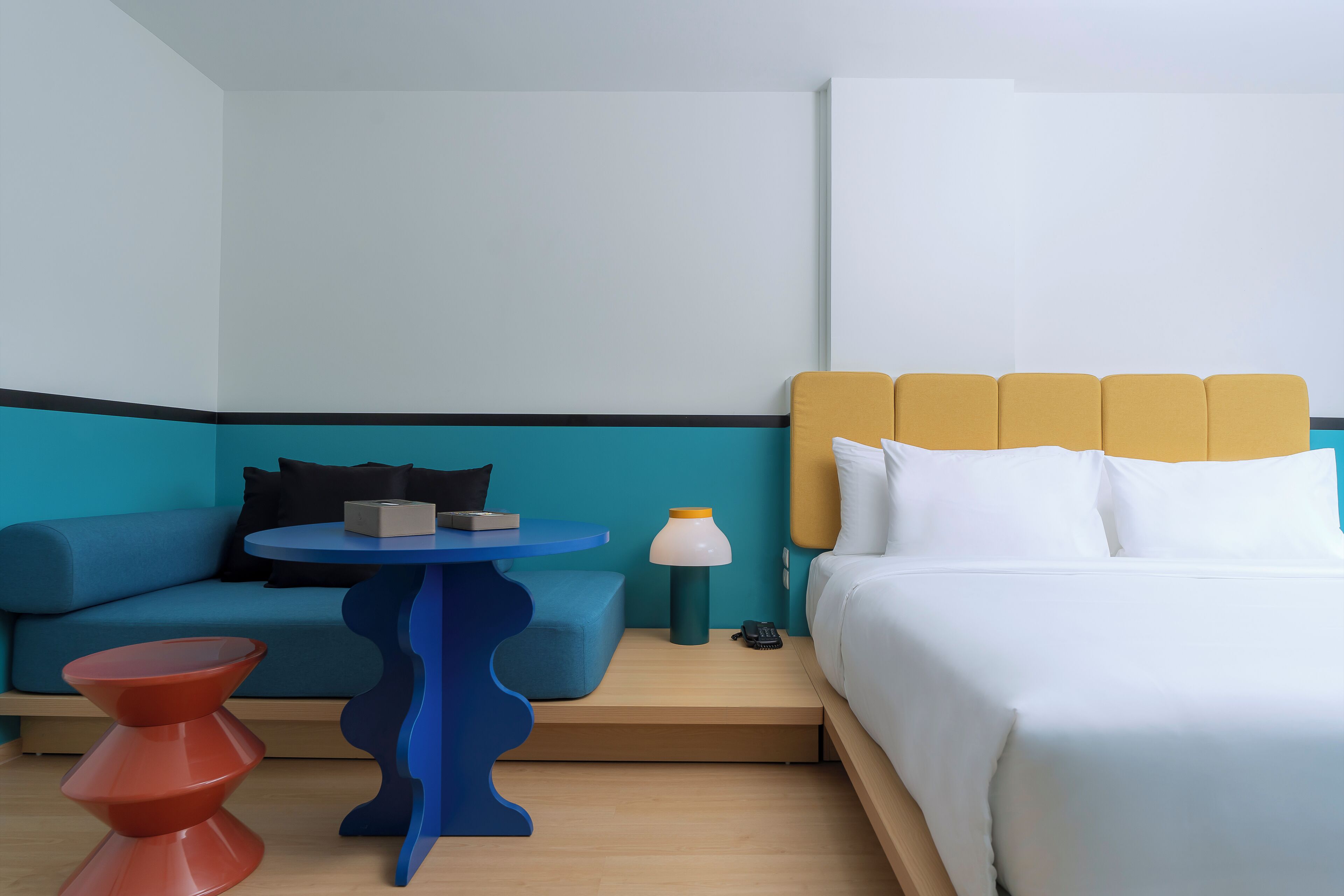 A modern hotel room with a blue sofa, a small round table, a red stool, a bed with white linens, and a yellow headboard.