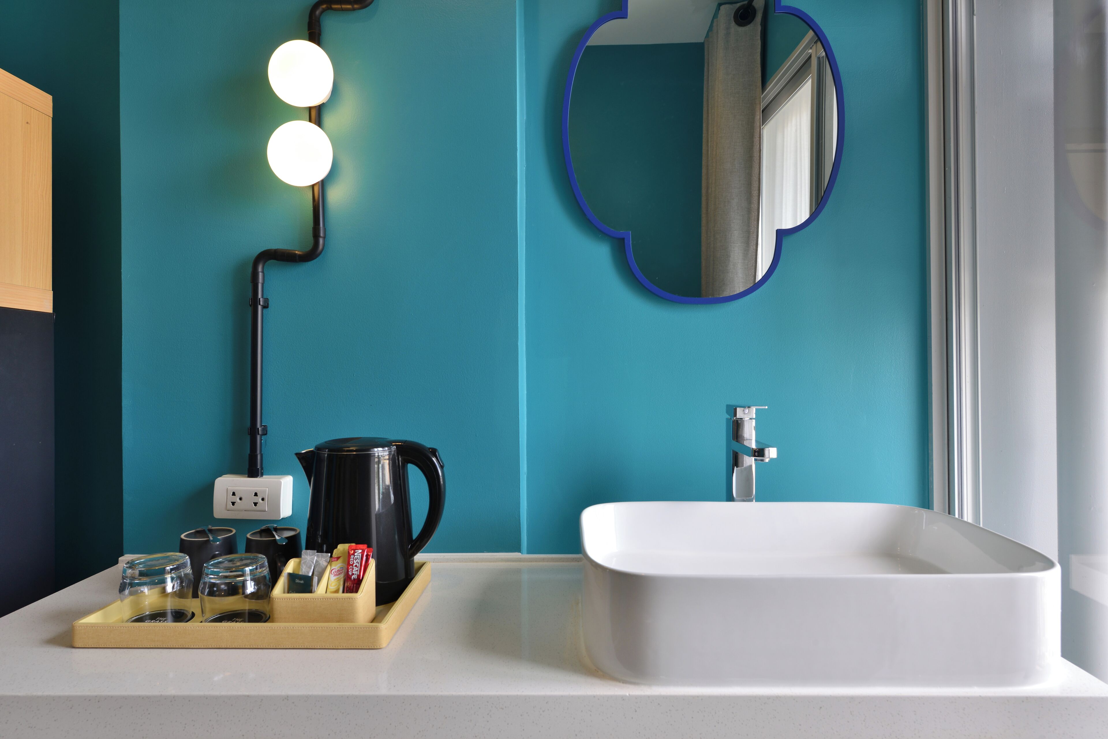A modern bathroom with a teal wall, a white sink, and a mirror with a blue frame. There are lights above the mirror, a black kettle, and a tray with glasses and a bottle.