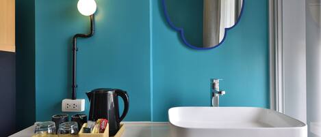 Un baño moderno con una pared verde azulado, un lavamanos blanco y un espejo con marco azul. Hay luces sobre el espejo, una tetera negra y una bandeja con vasos y una botella.