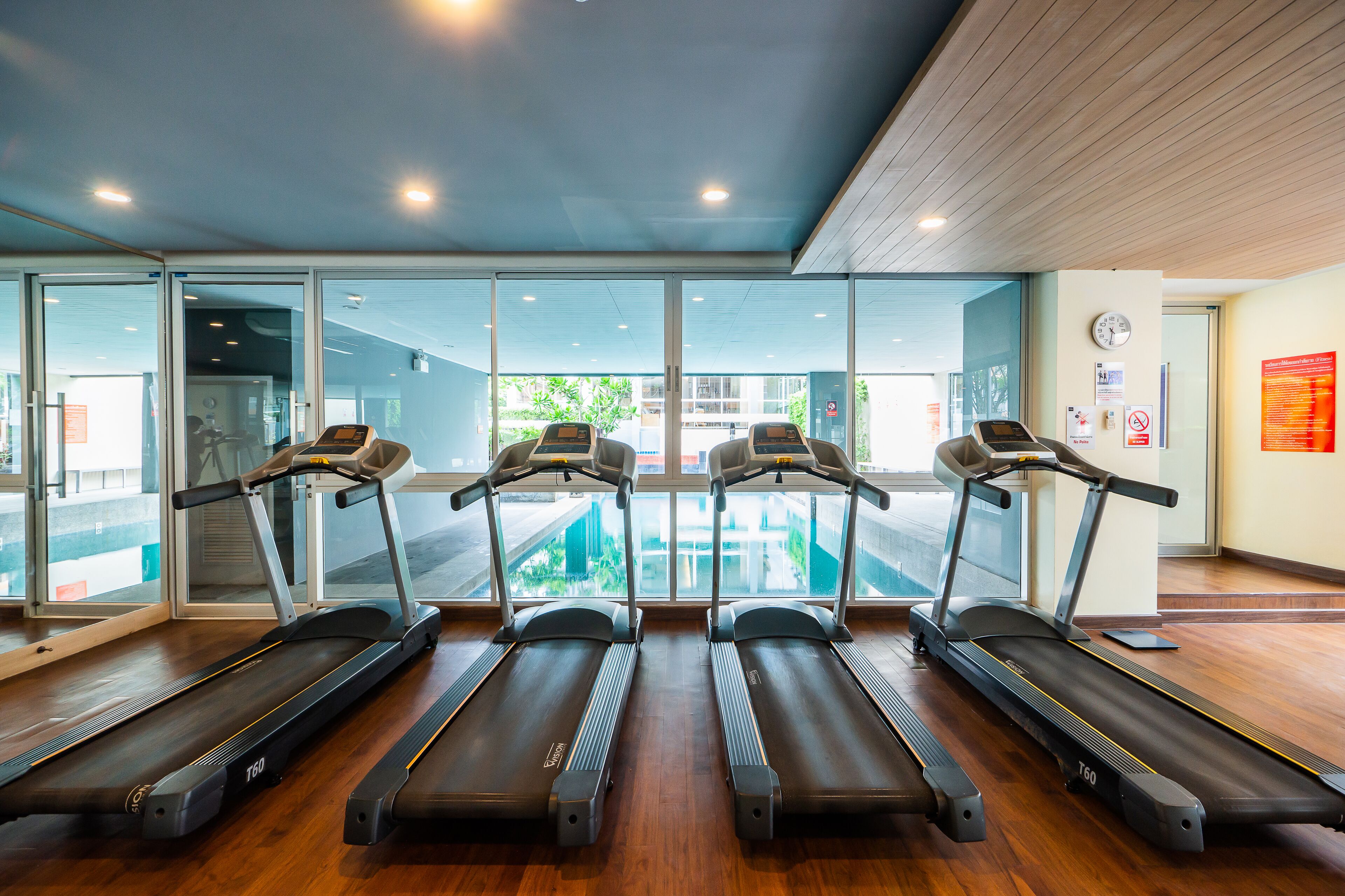 A gym with four treadmills aligned in a row, large windows, and a wooden floor.