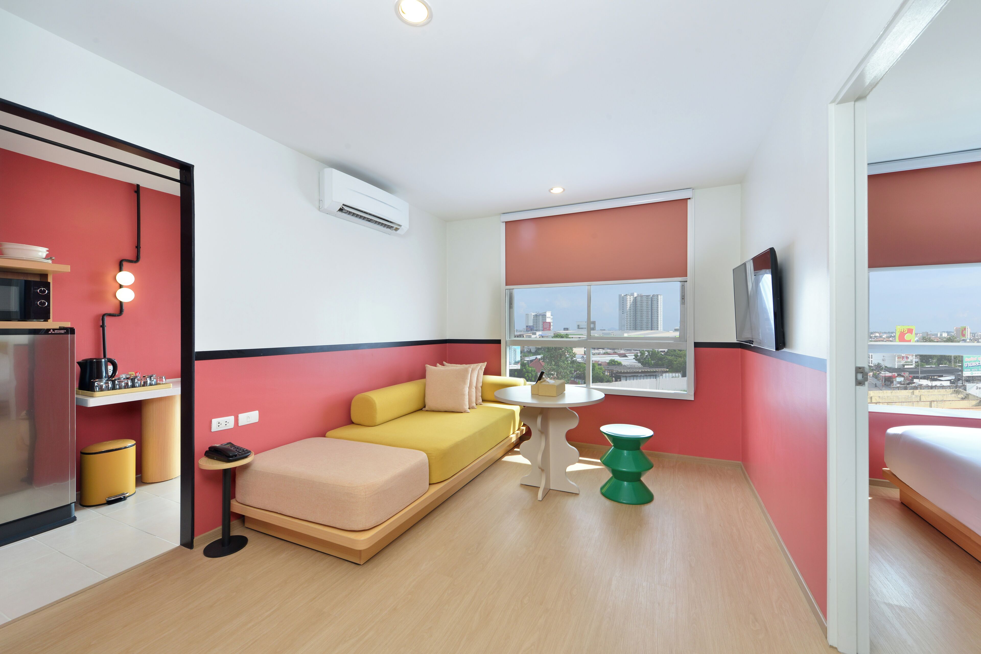 A compact hotel room with a sofa bed, a small kitchenette, a dining table, and a window with a city view.