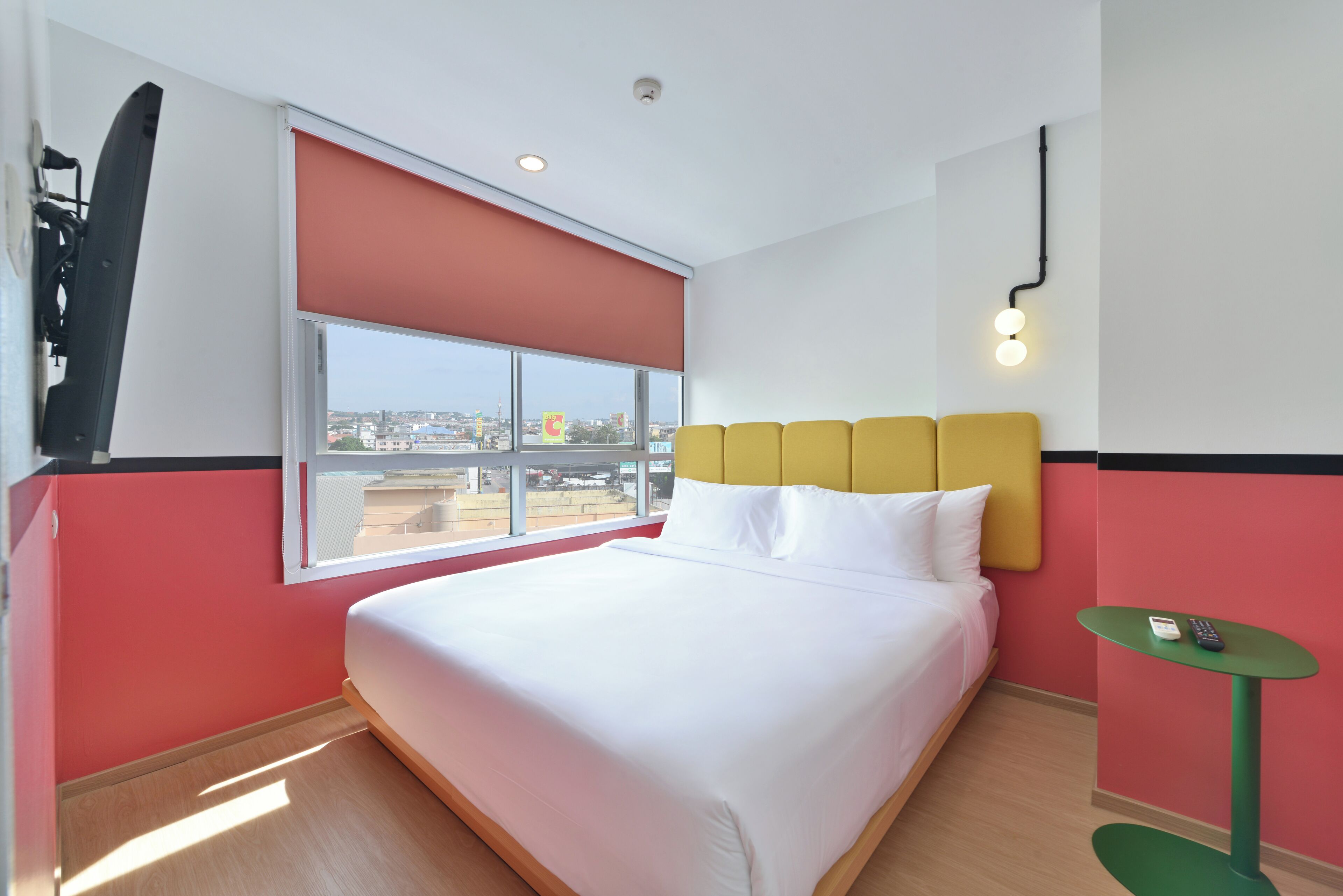 A hotel room with a bed, a window with a red blind, a green side table, and a city view.