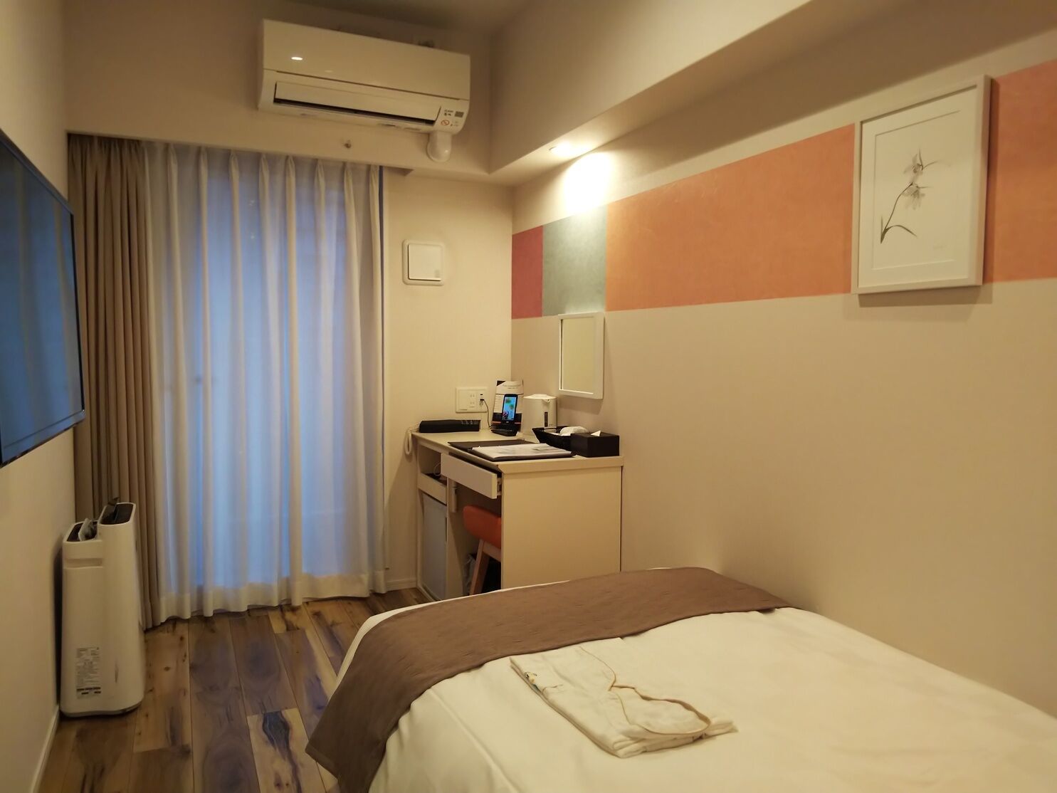 A hotel room with a bed, a desk with a chair, an air conditioning unit, a window with curtains, and a wall-mounted mirror.