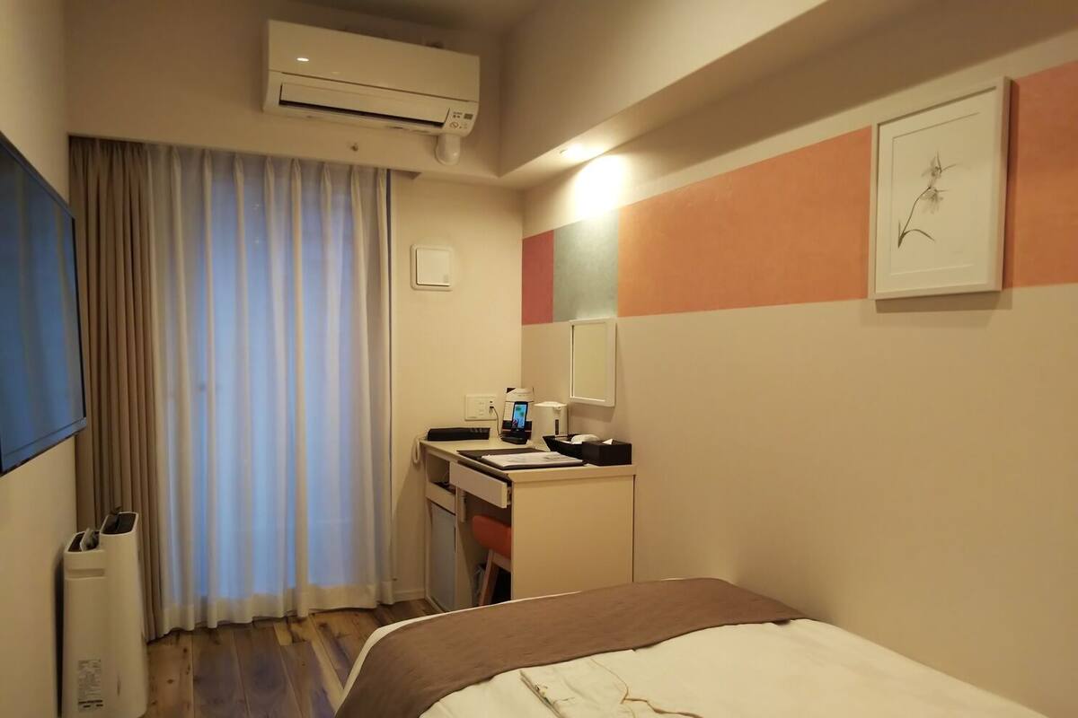 Ein Hotelzimmer mit einem Bett, einem Schreibtisch mit Stuhl, einer Klimaanlage, einem Fenster mit Vorhängen und einem wandmontierten Spiegel.