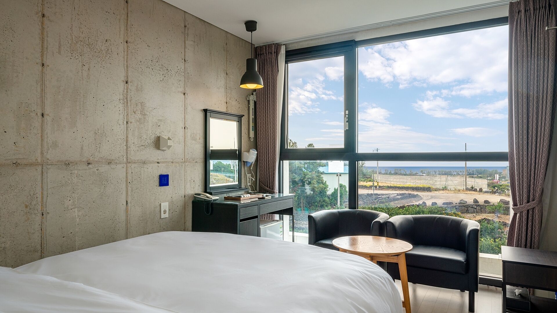 Quarto de hotel com uma cama grande, uma escrivaninha, uma cadeira e vista do horizonte através de grandes janelas.