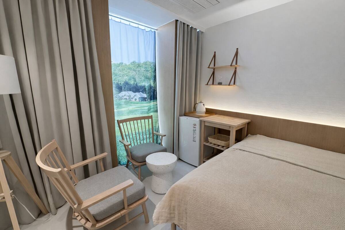 A hotel room with a bed, a chair, a small table, a mini-fridge, and a view of a green landscape through a window.