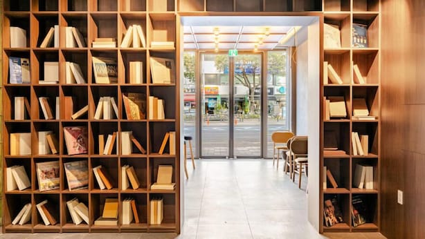 A bookstore with wooden shelves filled with books, a glass door entrance, and a seating area with chairs. - JEJU DSPHERE HOTEL (Jeju City)