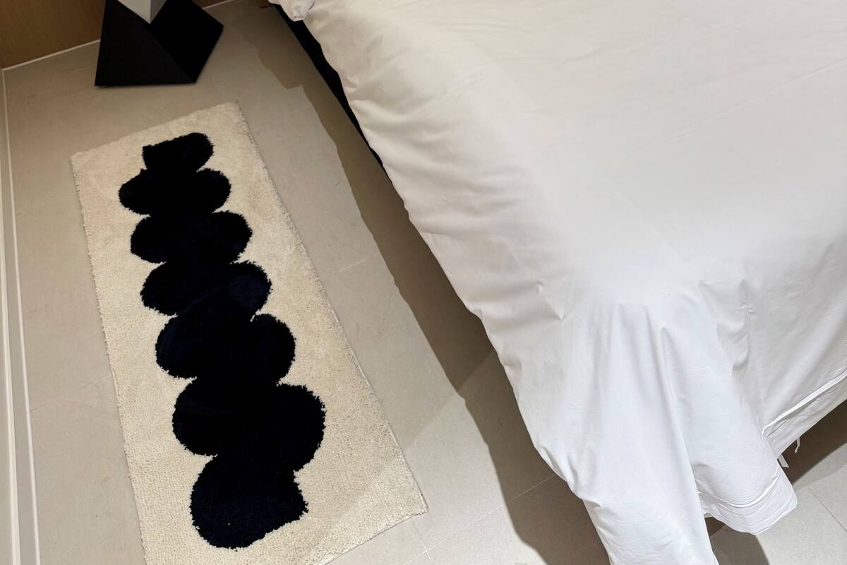 A neatly made bed with white bedding, green striped pillows, a white bedside table with a lamp, and a black and white rug.
