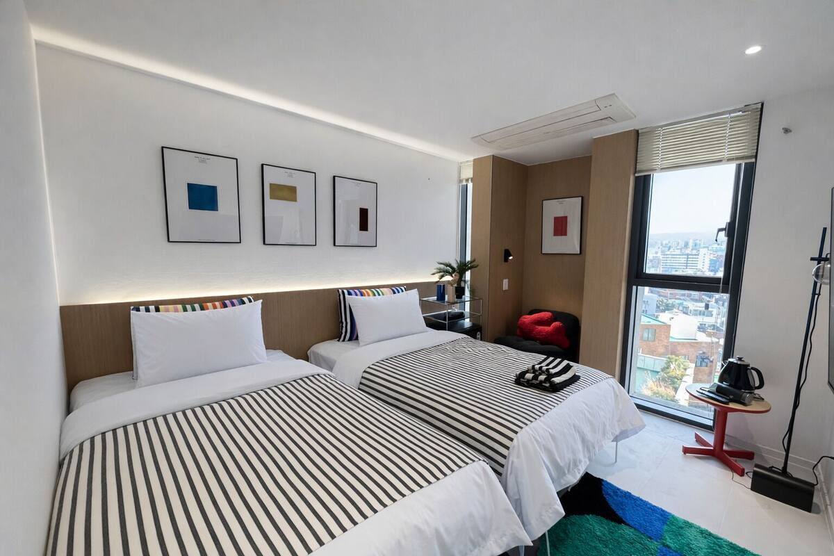 A hotel room with two beds, a large window with a city view, a small table with a kettle, and framed pictures on the wall.