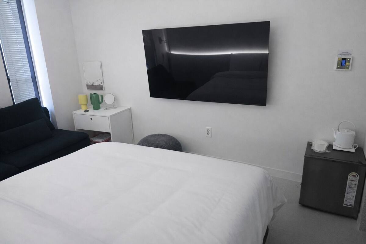 A hotel room with a bed, a TV mounted on the wall, a small bedside table with a lamp, and a nightstand with a kettle.