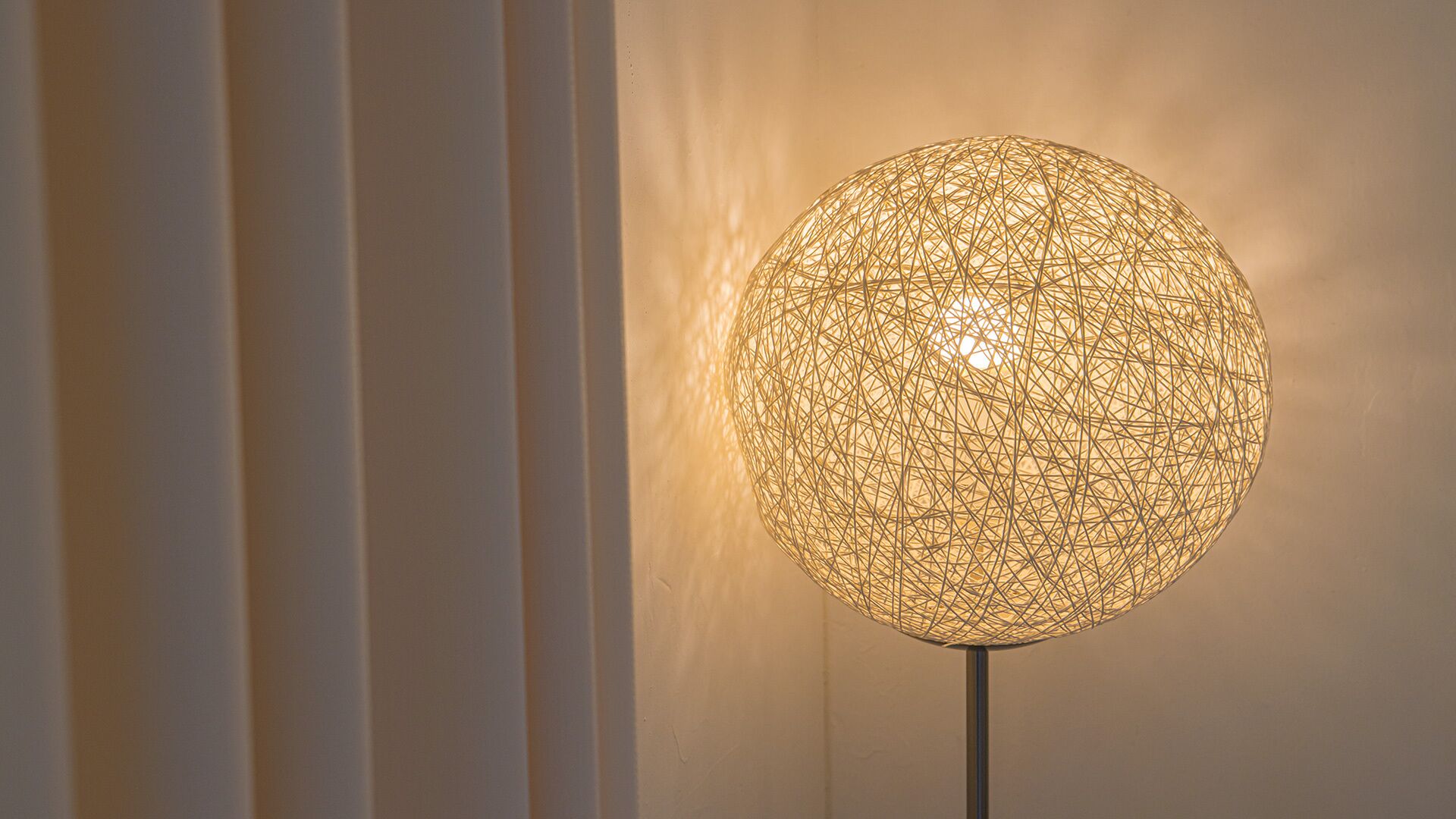 Una lámpara esférica de estructura metálica con una luz cálida, sobre un pie de lámpara en un pared lisa.