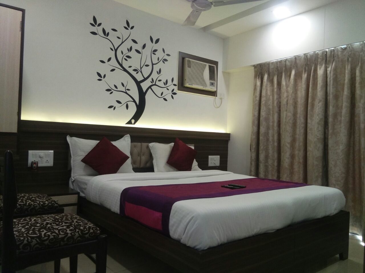 A hotel room with a bed, a wall-mounted air conditioner, a ceiling fan, and a window with curtains.