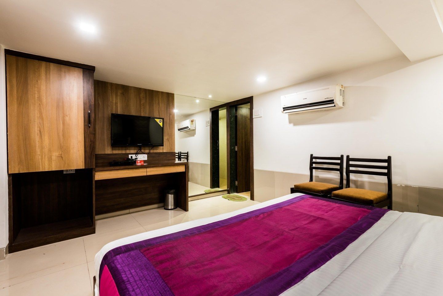 A hotel room with a bed, a desk with a TV, a chair, and an air conditioning unit.