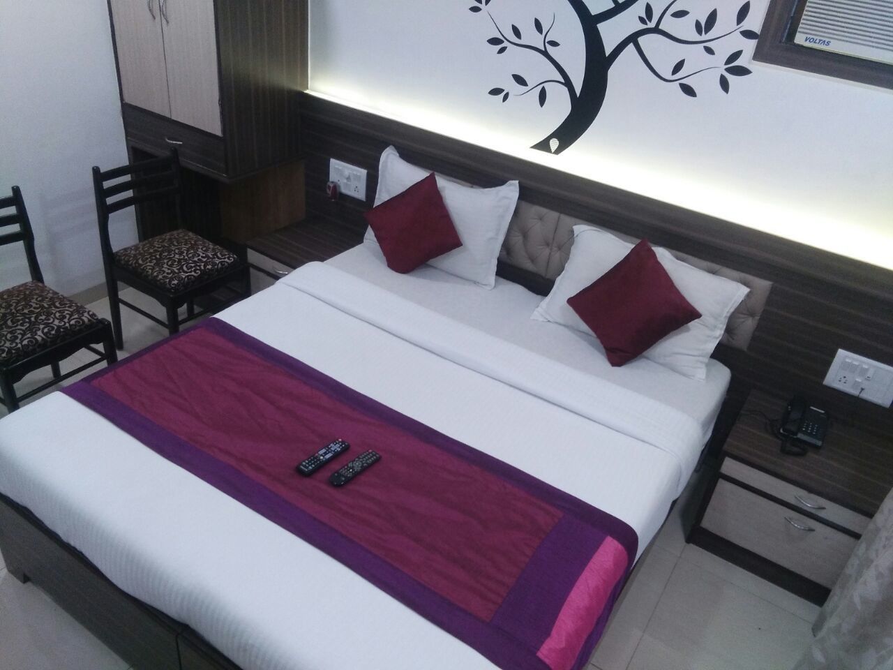 A neatly made bed with a red and white bedspread, two pillows, and two remote controls on the bed. There are two chairs with patterned cushions, a wall with a tree decal, and a nightstand with a phone.