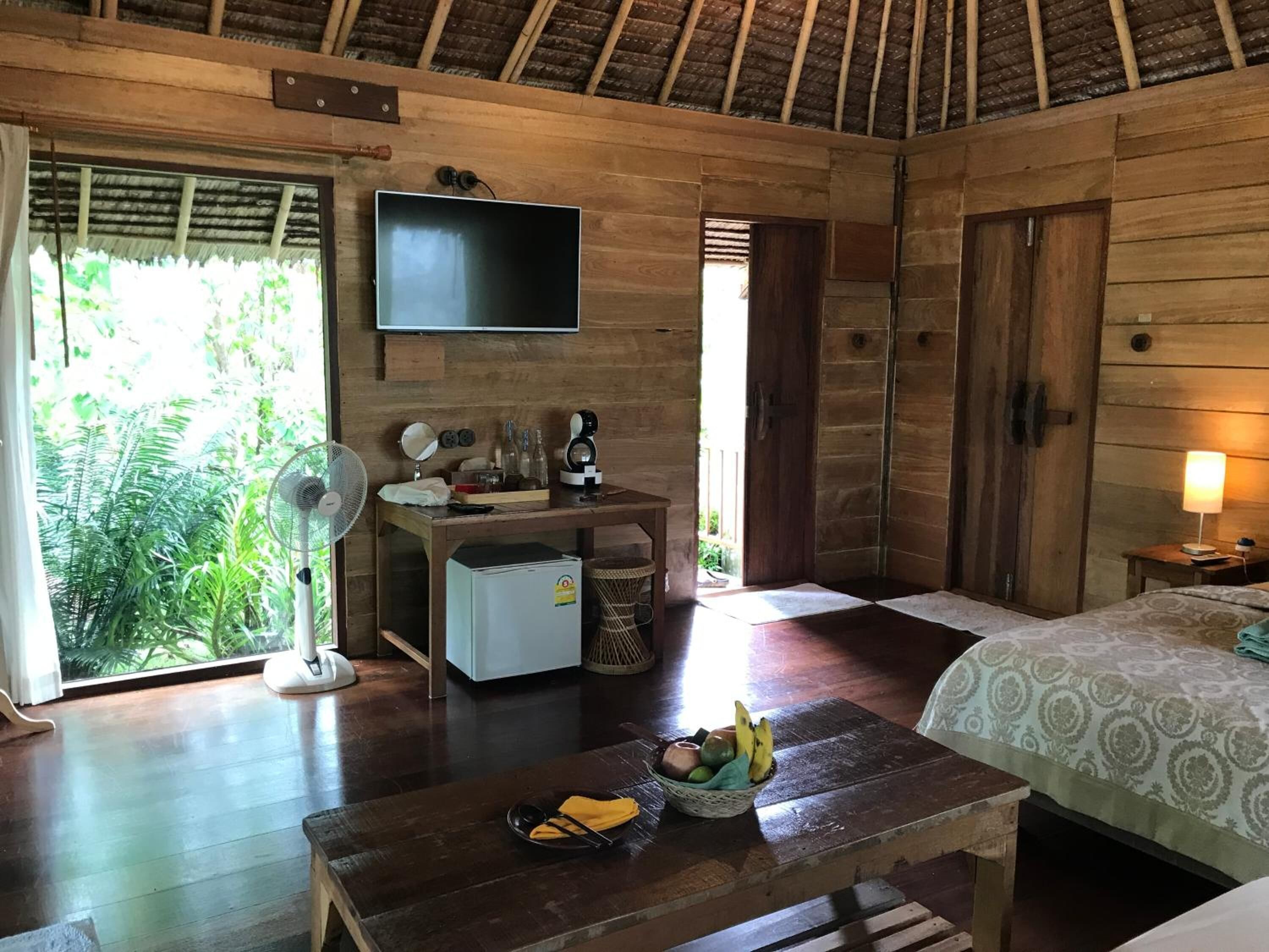 A wooden room with a bed, a desk with a TV, a fan, and a table with fruits.