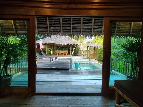 Una terraza de madera que conduce a una piscina con estructuras de techo de paja y asientos al aire libre.