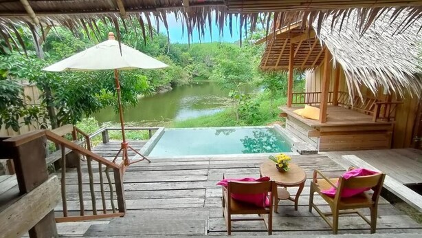 Terraza de madera con piscina, sillas y sombrilla, con vistas a un paisaje natural.
