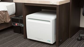 A compact, white refrigerator under a wooden counter in a hotel room, with a black trash bin and a bed with a brown blanket visible.