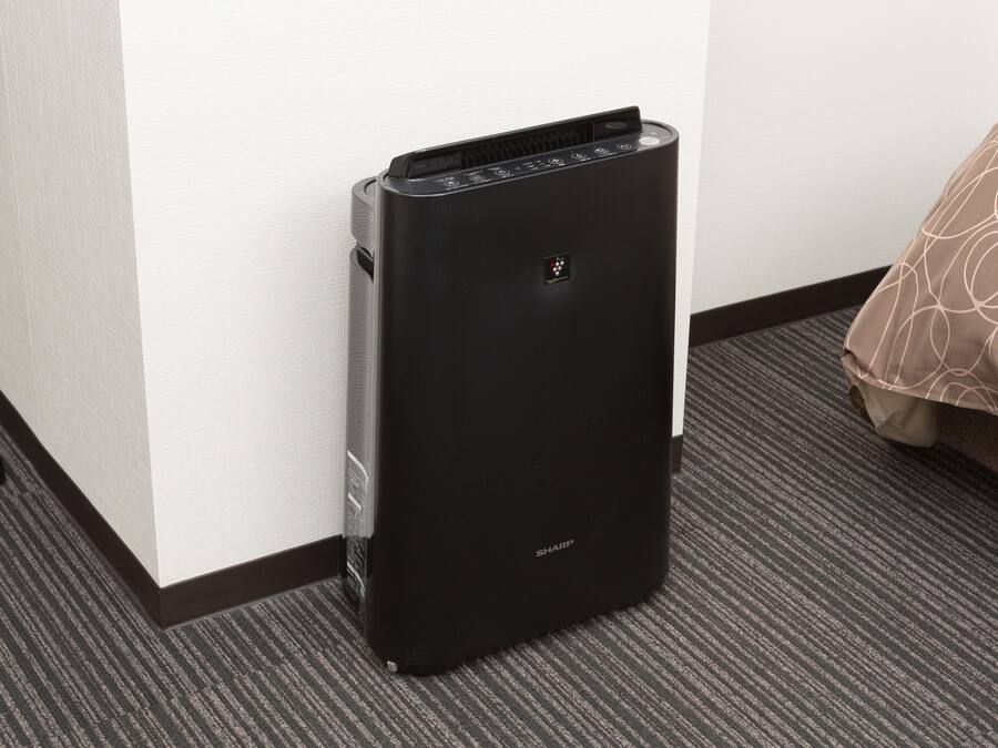 Um aquecedor portátil preto com painel de controle, posicionado contra uma parede em quarto de hotel com carpete e colcha estampada.