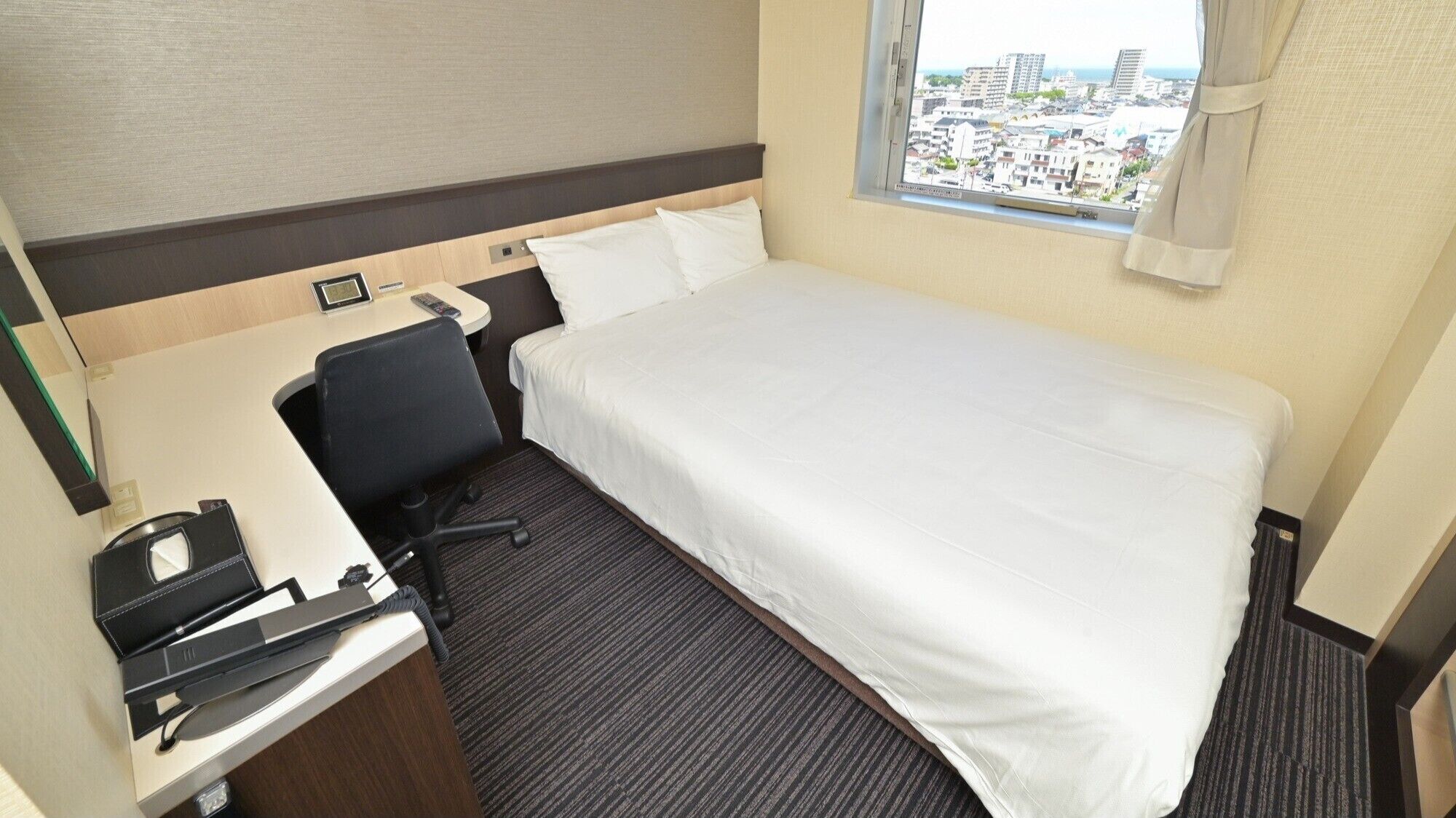 Habitación de hotel con cama, escritorio con silla, ventana con vista a edificios y piso alfombrado.