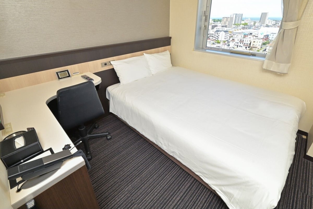 A hotel room with a bed, a desk with a chair, a window with a view of buildings, and a carpeted floor.