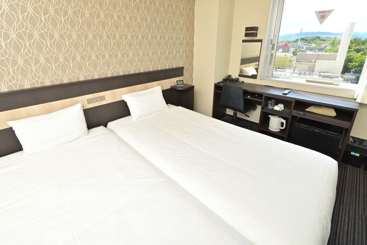 A hotel room with a large bed, bedside tables, a desk with a chair, a mirror, and a window with a view.