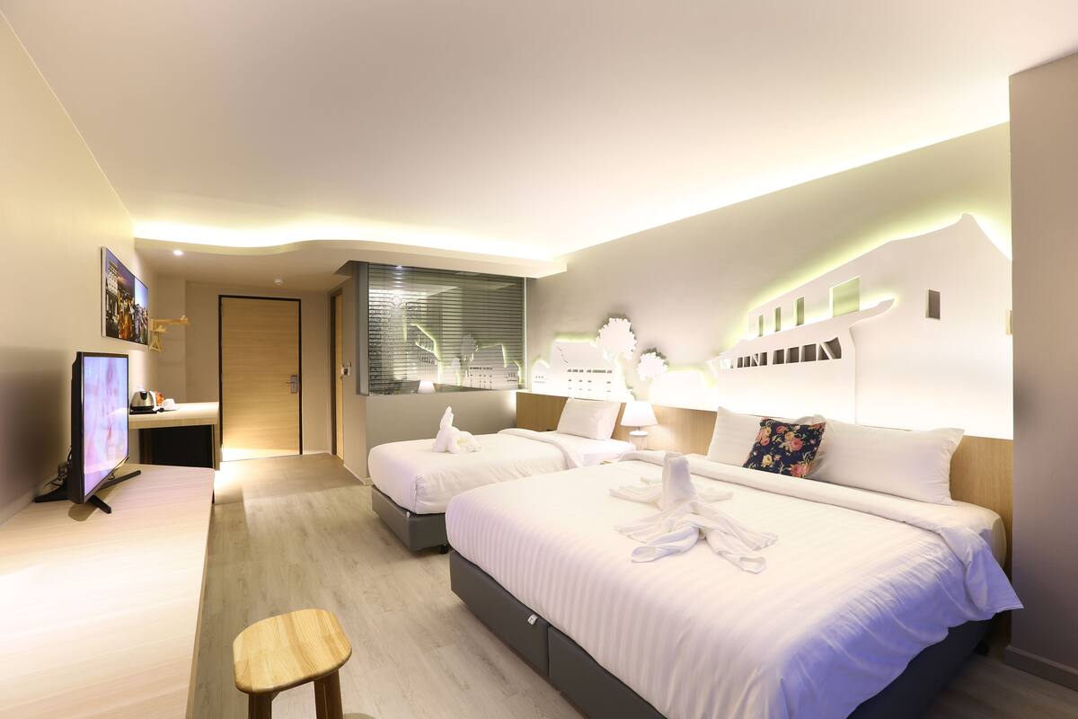 A modern hotel room with two beds, a desk, a television, and a window with blinds.
