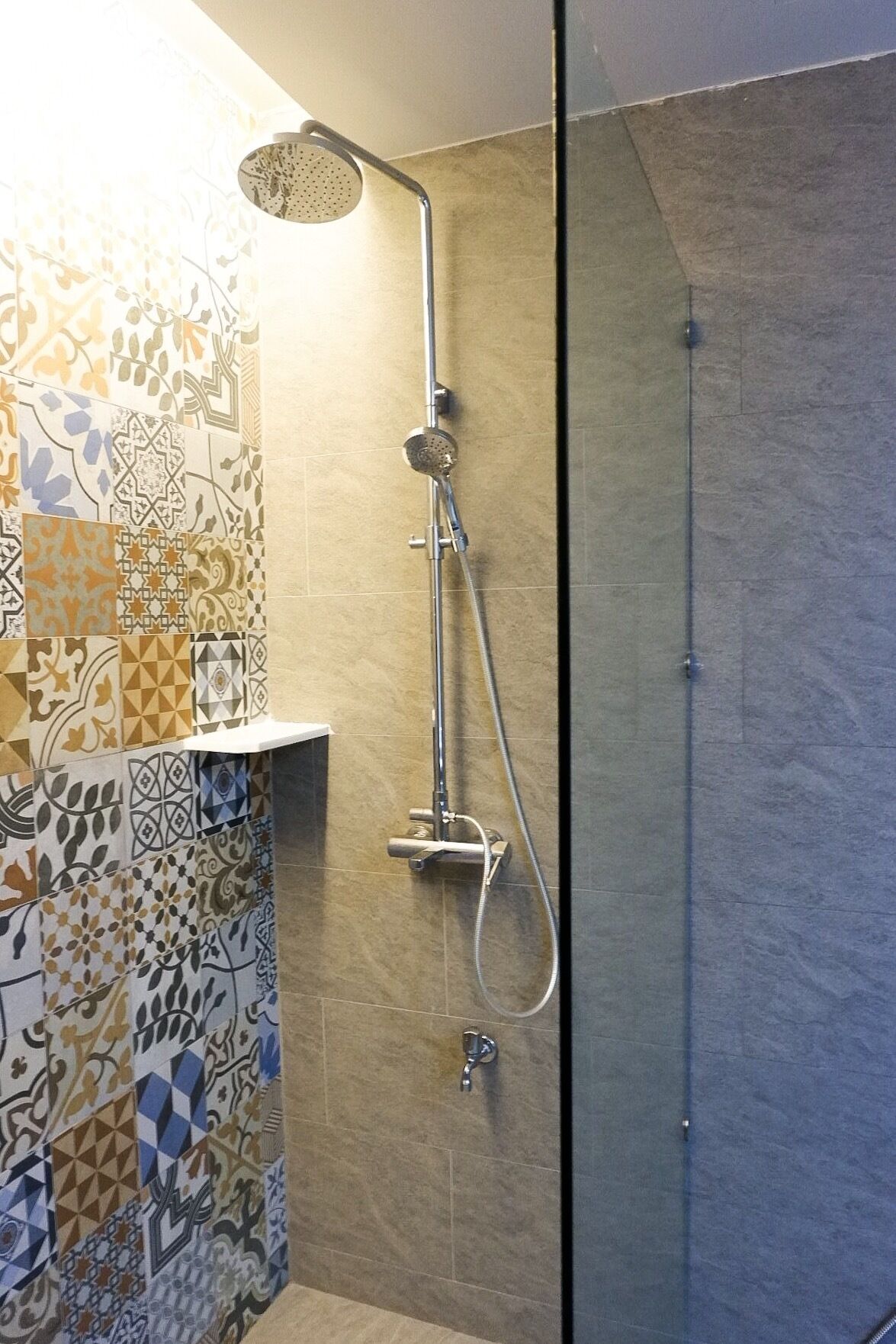 A shower with a handheld showerhead and a wall-mounted faucet, set against a tiled wall with a geometric pattern.