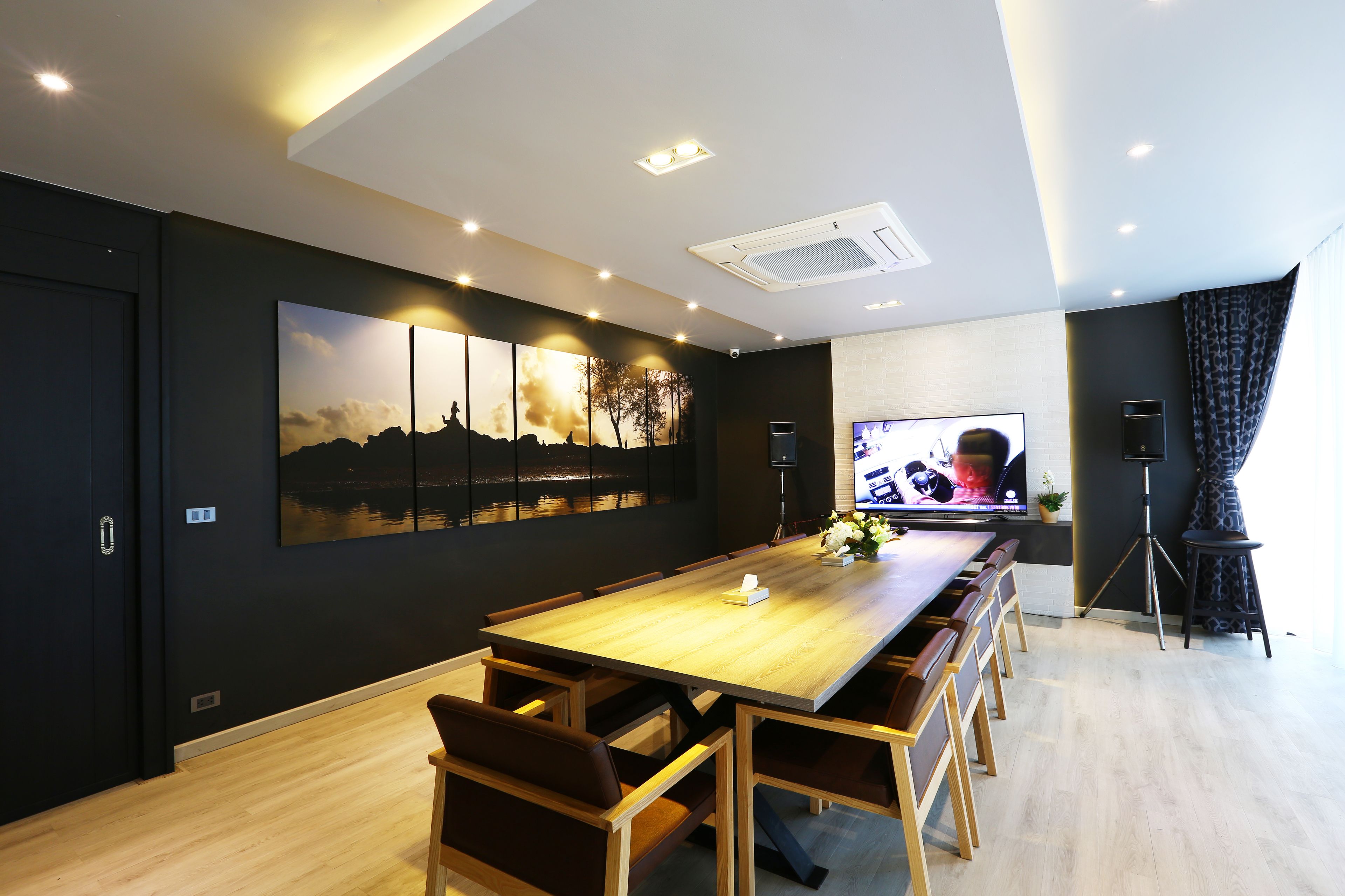 A modern dining area with a long wooden table, multiple chairs, a large wall-mounted TV, and a ceiling-mounted air conditioning unit.