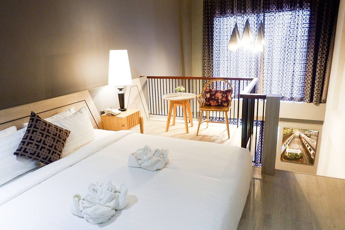 A hotel room with a bed, a nightstand, a lamp, a chair, a small table, and a balcony with patterned curtains.