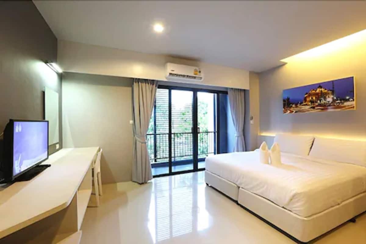A hotel room with a bed, a desk with a computer, an air conditioning unit, and a large window with curtains.