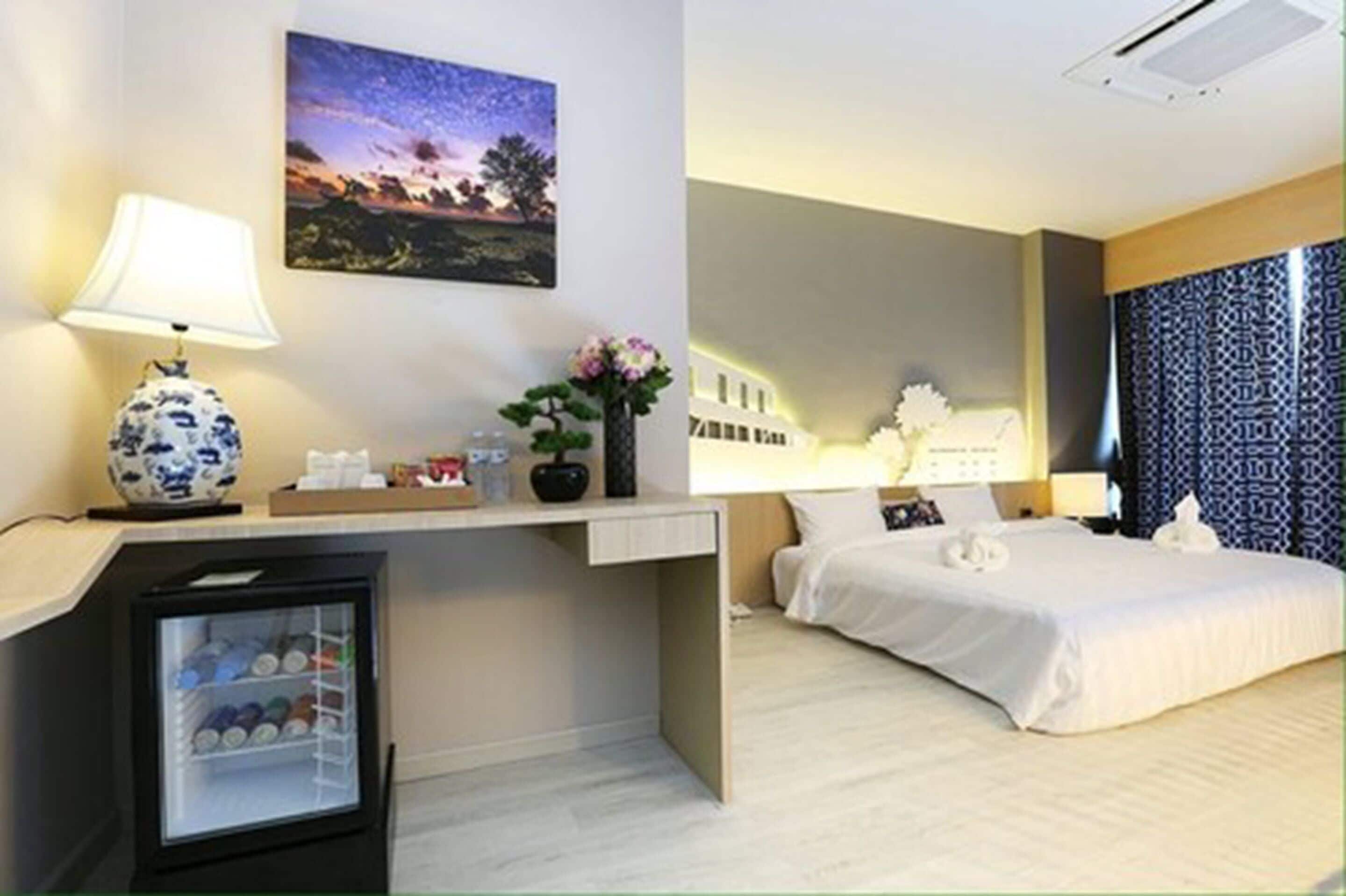 Habitación de hotel con cama, escritorio con lámpara, refrigerador pequeño, mesita de noche y un cuadro colgado en la pared.