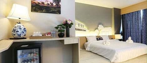Habitación de hotel con cama, escritorio con lámpara, refrigerador pequeño, mesita de noche y un cuadro colgado en la pared.
