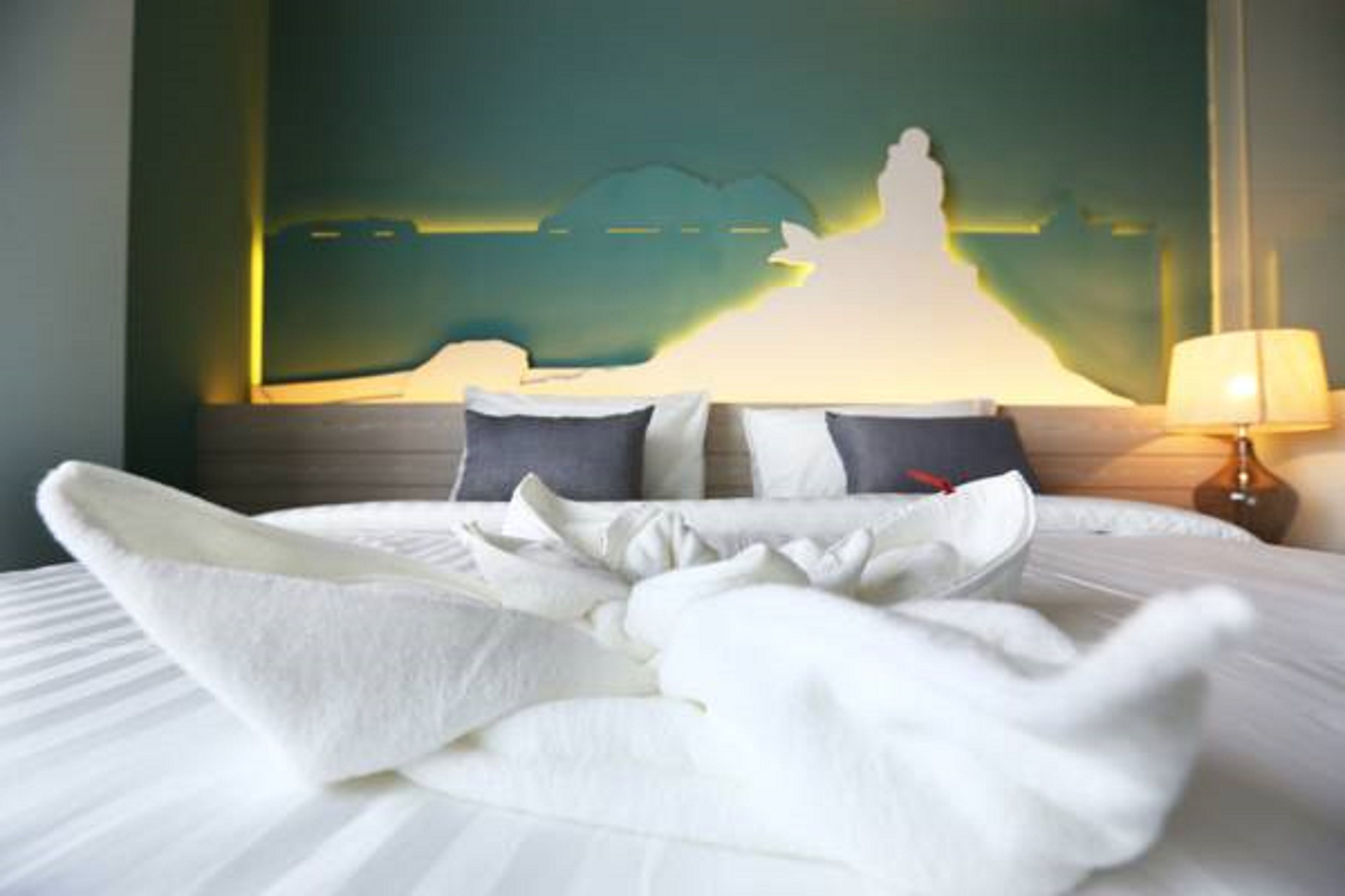 A neatly made bed with white linens and a folded white towel, a lamp on the bedside table, and a wall with a silhouette artwork.